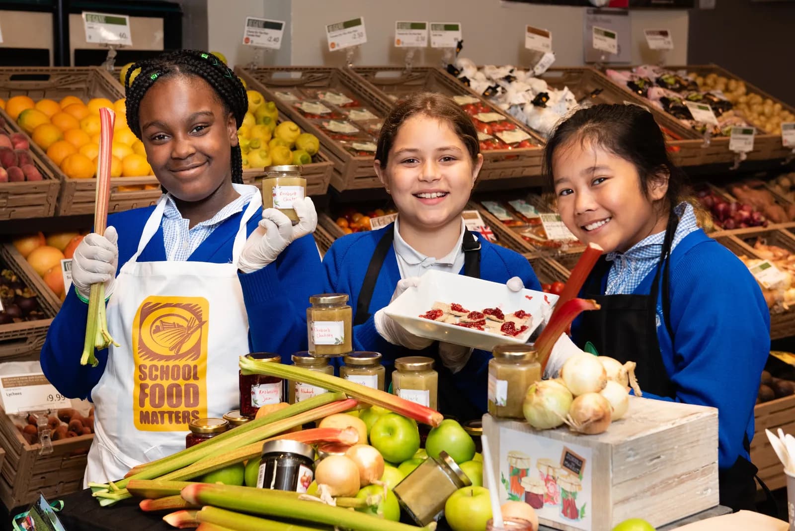 Three smiling students in aprons stand behind a table laden with fresh produce and jars of preserves, in what appears to be a grocery store.